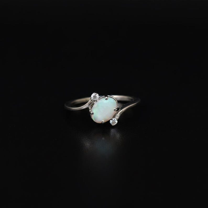 May include: A silver ring featuring an oval opal gemstone and two smaller accent stones. The opal has a milky white appearance. The ring's band curves gracefully around the main stone, with the accent stones set on either side.