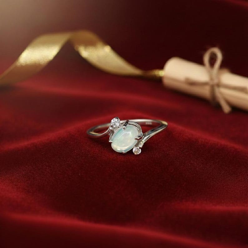 May include: A silver ring with an oval, opalescent gemstone and two smaller accent stones. The ring sits on a red velvet surface, with a gold ribbon and a rolled scroll in the background.