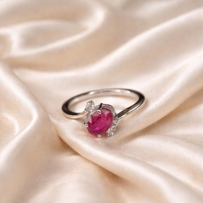May include: A silver ring with a large, oval-cut ruby gemstone as the centerpiece. The ruby is a deep, vibrant red. Small, clear gemstones are set on either side of the ruby. The ring is displayed on a cream-colored, satin-like fabric.