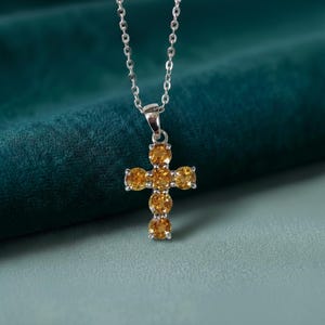 Peut inclure: Un collier en argent avec un pendentif en forme de croix orné de pierres précieuses rondes jaune doré. La croix est composée de cinq pierres précieuses verticalement et quatre horizontalement, créant un bijou symétrique et élégant.