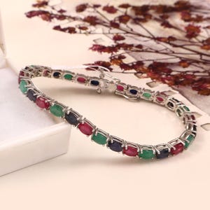 Puede incluir: Una pulsera de plata con piedras preciosas ovaladas alternas en rojo, verde y azul. La pulsera se presenta sobre un fondo suave y neutro con elementos florales.