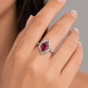 Könnte beinhalten: Ein silberfarbener Ring mit einem zentralen, oval geschliffenen, tiefroten Rubin. Der Rubin ist von einer marquiseförmigen Fassung umgeben, die mit kleinen, klaren, rund geschliffenen weißen Steinen besetzt ist, die sich entlang der Ringschiene fortsetzen.
