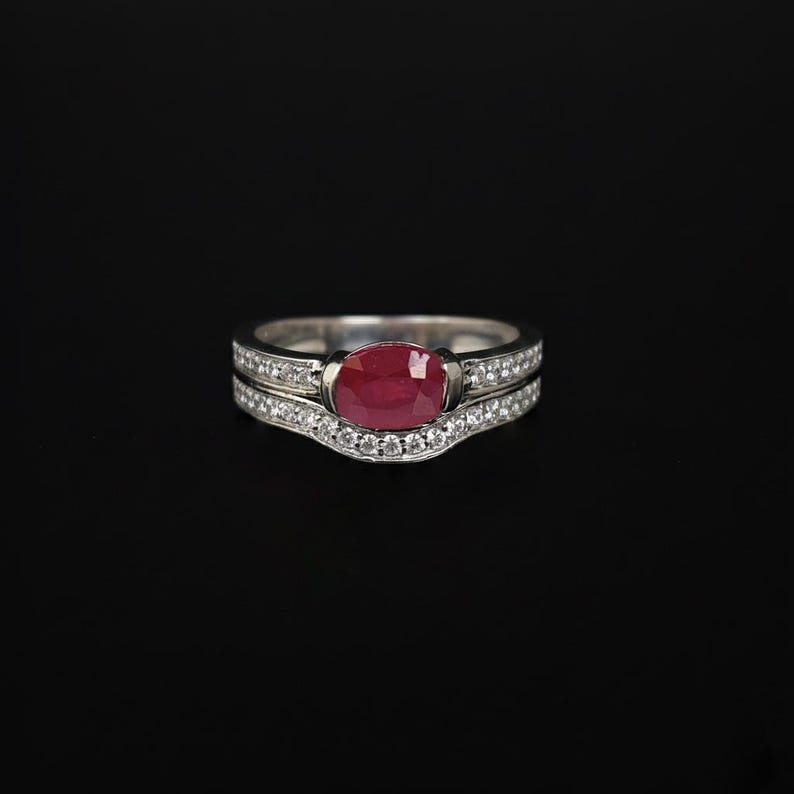 This exquisite bridal set features a vibrant, oval-cut ruby nestled within a shimmering halo of pavé-set diamonds. Crafted in high-quality white gold, the contoured wedding band fits seamlessly against the engagement ring, creating a sophisticated, layered look. Whether you are celebrating a July birthday, a 40th anniversary, or a romantic proposal, this set offers a bold pop of color and classic elegance that stands out from the crowd.