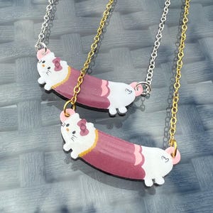May include: Two necklaces with a cartoon cat pendant. The pendants are white with a pink and maroon body, and a pink bow. One necklace has a silver chain, the other a gold chain.