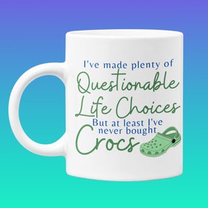 May include: White ceramic coffee mug with a green and blue design. The mug reads "I've made plenty of Questionable Life Choices But at least I've never bought Crocs".