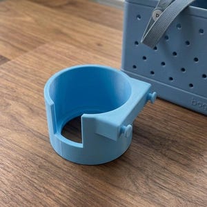 May include: A light blue cup holder attachment with a cylindrical body and two small protruding pegs on the side for mounting. The cup holder features a cutout section on the front rim and is positioned next to a light blue perforated tote bag.