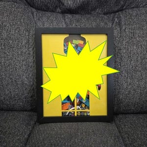 May include: A framed artwork with a black frame and a yellow background. The artwork features a collection of colorful objects, partially obscured by a large yellow starburst shape. The frame is rectangular and sits on a gray surface.