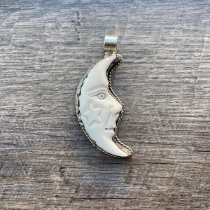 May include: A white crescent moon pendant with a carved face and stars. The pendant is made of bone and has a gold-tone metal trim.