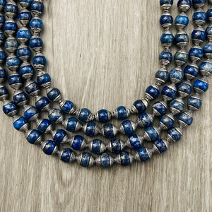 Puede incluir: Un collar de múltiples hilos hecho de cuentas de lapislázuli azul oscuro con detalles plateados. Las cuentas son redondas y tienen una forma ligeramente irregular. El collar está colocado sobre una superficie de madera.