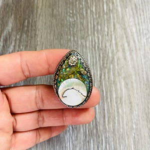 May include: A teardrop-shaped silver ring with a mosaic of green, gold, and iridescent abalone shell. The ring features a white, crescent-shaped inlay and a decorative floral accent at the top. The ring is held in a hand.