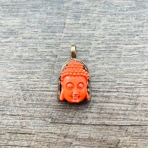 May include: A pendant featuring a detailed orange resin Buddha head with a gold-toned metal frame.