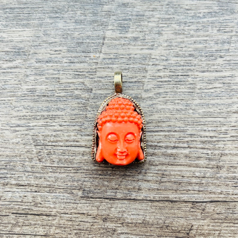 May include: A pendant featuring a detailed orange resin Buddha head with a gold-toned metal frame.