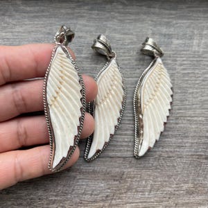 May include: Three pendant necklaces with a wing design. Each pendant features a cream-colored wing shape with detailed feathering, encased in a silver-toned metal frame. The pendants are displayed on a wooden surface.