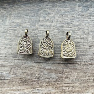 May include: Three ornate gold-toned metal pendants with intricate floral designs. The pendants are shaped like small bells and have a loop at the top for attaching to a chain or cord.