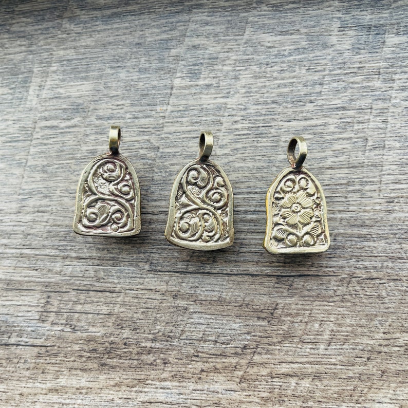 May include: Three ornate gold-toned metal pendants with intricate floral designs. The pendants are shaped like small bells and have a loop at the top for attaching to a chain or cord.