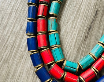 May include: Close-up of three strands of colorful beaded necklaces. The beads are cylindrical with gold-colored metal accents. The colors include blue, red, and turquoise. The necklaces are arranged in a curved pattern against a gray wooden surface.