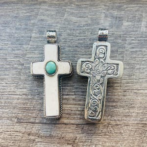 Puede incluir: Dos colgantes de cruz plateados. Una cruz tiene un centro blanco con una piedra turquesa y un borde plateado. La otra cruz está grabada con motivos florales. Ambos tienen lazos decorativos para colgar.
