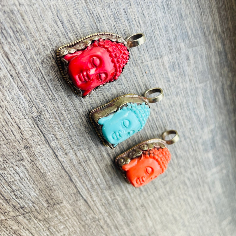 May include: Three silver and coral Buddha head pendants. The pendants are in the shape of a Buddha head with a closed eye and a serene expression. The pendants are on a wooden surface.