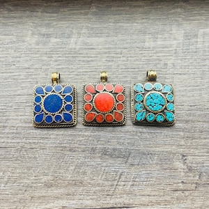 May include: Three square pendants with brass-colored metal frames. Each pendant features a central stone surrounded by smaller stones in a square pattern. The stones are blue, red, and turquoise. The pendants are displayed on a wooden surface.