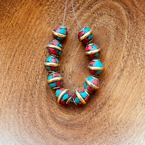 Puede incluir: Una hebra de cuentas redondas con un patrón de mosaico turquesa y rojo. Las cuentas están enhebradas en un hilo transparente y tienen un borde dorado.