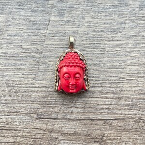 May include: A red coral Buddha pendant with a silver frame. The pendant is detailed with a carved face and a textured surface.