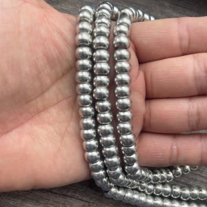May include: Strands of silver-colored beads, each with a rounded shape, are held in a person's hand. The beads are strung together, forming multiple rows. The beads have a metallic sheen and reflect light.