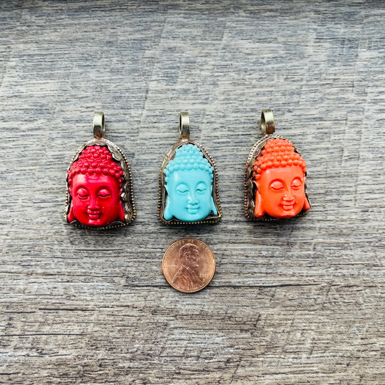 May include: Three Buddha head pendants with different colored stone inlays. The pendants are red, blue, and orange. The pendants are on a wooden surface.