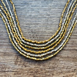 May include: Close-up of multiple strands of beaded necklaces. The necklaces feature a mix of gold and silver-colored faceted beads. The beads are strung together, creating a layered effect. The background is a wooden surface.