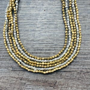 Puede incluir: Un collar de múltiples hilos con filas alternas de cuentas cuadradas doradas y plateadas. El collar se exhibe sobre una superficie de madera, mostrando su diseño en capas y brillo metálico. Esta joya ofrece una mezcla de elegancia y simplicidad.