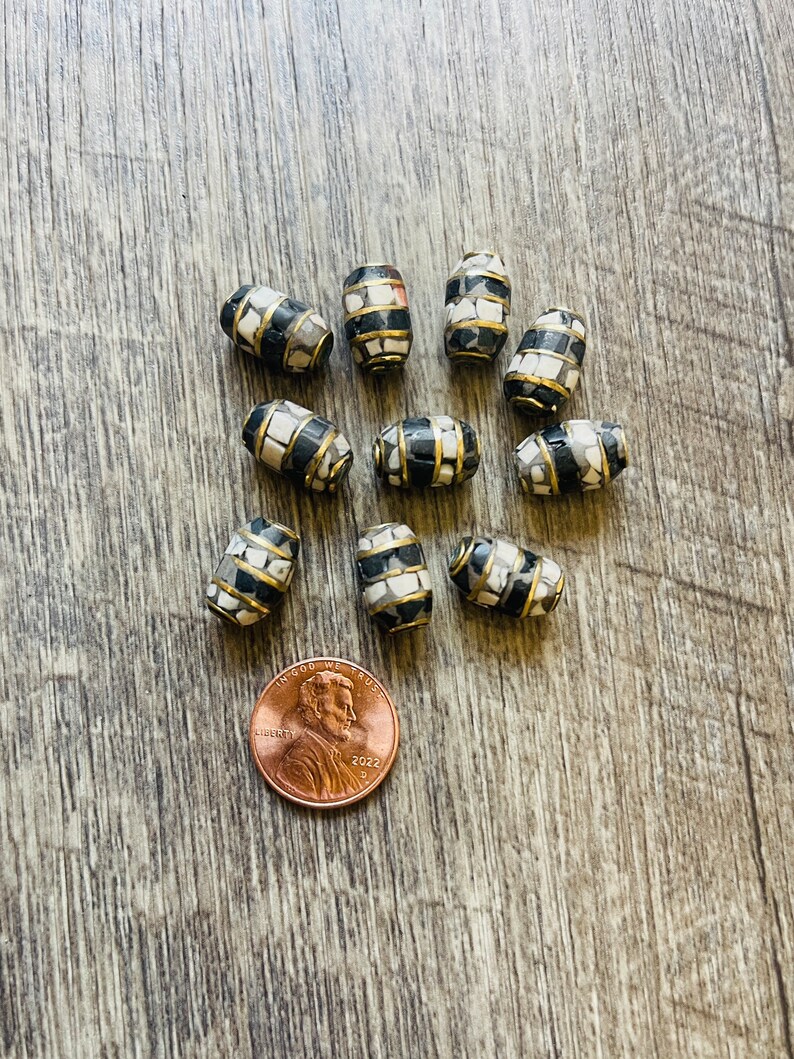 May include: A collection of 10 black and white mosaic barrel-shaped beads with gold trim. The beads are approximately the size of a US penny.