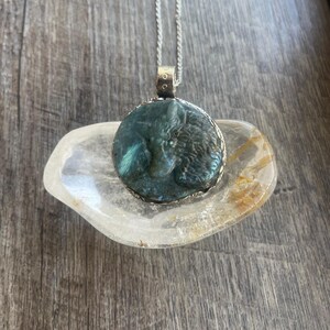Pode incluir: Um colar de corrente de prata com um pingente redondo que apresenta uma pedra de labradorita esculpida. A pedra é de cor azul-esverdeada escura com brilhos iridescentes.