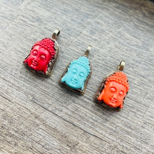 May include: Three pendants featuring Buddha faces carved from red, blue, and orange stone. The pendants are set in silver-toned metal frames.