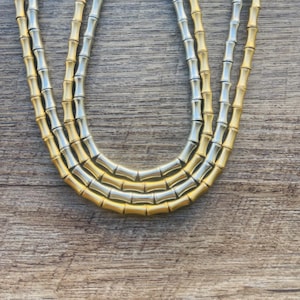 May include: A layered necklace with a bamboo design. The necklace features alternating gold and silver-colored segments, creating a two-tone effect. The jewelry is displayed on a wooden surface.