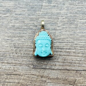 May include: A turquoise blue Buddha pendant with a gold-toned frame. The pendant is a detailed depiction of the Buddha's face.