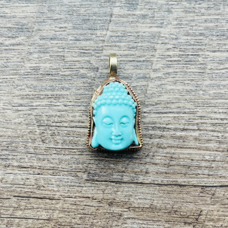 May include: A turquoise blue Buddha pendant with a gold-toned frame. The pendant is a detailed depiction of the Buddha's face.