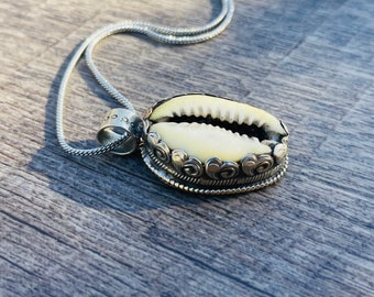 May include: A silver chain necklace with a cowrie shell pendant. The shell is surrounded by a silver frame with intricate designs.