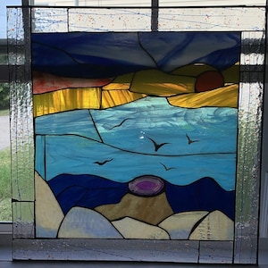 May include: A stained glass window depicting a sunset over a body of water with birds flying overhead. The sky is a gradient of blue, purple, and orange. The water is a light blue with a hint of green. The birds are black silhouettes against the sky. The window is framed in a dark wood frame.