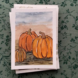Könnte beinhalten: Eine rechteckige Grußkarte mit einem Aquarell von drei orangefarbenen Kürbissen mit braunen Stielen, die auf einer dunklen, strukturierten Oberfläche vor einem Hintergrund aus beigem und blauem Himmel ruhen. Die Karte ist weiß.