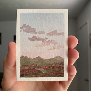 May include: A small rectangular watercolor painting on textured paper depicting a landscape at sunset. The scene features a pale blue and pink sky with soft, muted mauve clouds above a dark green and deep red textured horizon line.