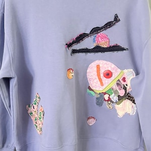 May include: A light purple crewneck sweatshirt featuring various fabric appliqués. The design includes a black jagged shape, a pink fish-like figure with a red button eye, a small floral patch, and other textured fabric scraps sewn onto the front.
