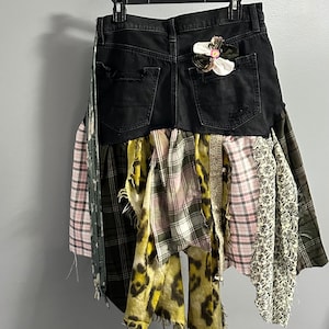 Puede incluir: Falda vaquera negra con detalles desgastados, adornada con un patchwork de varias telas. La falda presenta un adorno floral en el bolsillo trasero y una mezcla de telas de cuadros, florales y estampadas de animales.