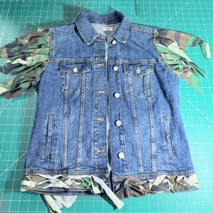 Puede incluir: Un chaleco de mezclilla azul con cuello cl&aacute;sico, cierre frontal de botones met&aacute;licos y dos bolsillos en el pecho. La prenda est&aacute; decorada con flecos de tela de camuflaje en los hombros y un borde trenzado de tela de camuflaje en el dobladillo.
