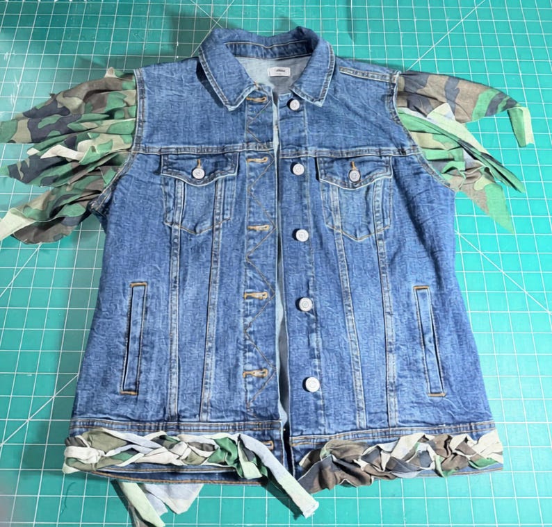 Puede incluir: Un chaleco de mezclilla azul con cuello cl&aacute;sico, cierre frontal de botones met&aacute;licos y dos bolsillos en el pecho. La prenda est&aacute; decorada con flecos de tela de camuflaje en los hombros y un borde trenzado de tela de camuflaje en el dobladillo.
