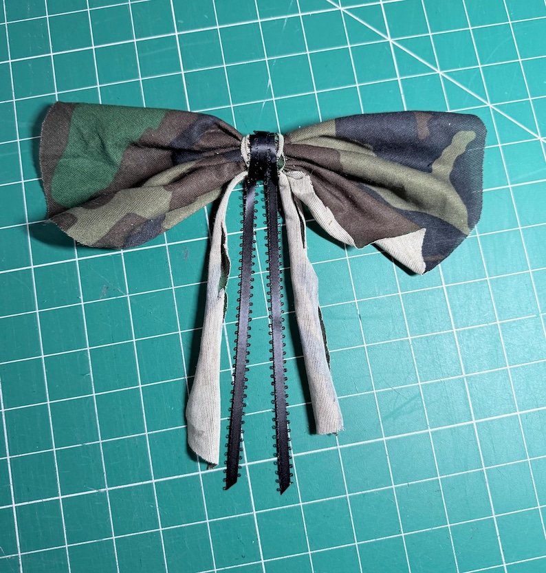 Puede incluir: Un lazo de tela con un patr&oacute;n de camuflaje en tonos verde, marr&oacute;n y negro. El centro est&aacute; envuelto en una cinta negra, con dos cintas negras largas y finas y dos tiras de tela que cuelgan del nudo del lazo.