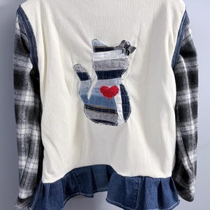 May include: Cream-colored corduroy jacket with a cat applique on the back. The cat is made of denim and striped fabric with a red heart. The sleeves are black and white plaid, and the bottom has a denim ruffle.