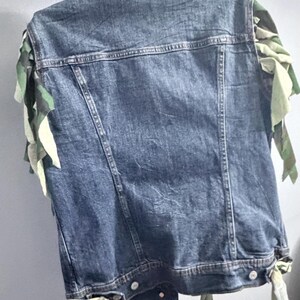 Puede incluir: Un chaleco de mezclilla azul con cuello cl&aacute;sico, costuras verticales y botones de metal en el dobladillo. Las sisas est&aacute;n adornadas con tiras de tela deshilachadas y superpuestas en un patr&oacute;n de camuflaje verde, marr&oacute;n y beige.