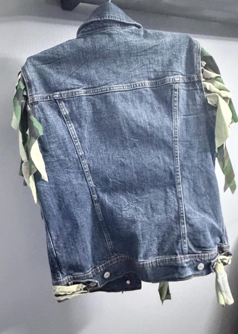 Puede incluir: Un chaleco de mezclilla azul con cuello cl&aacute;sico, costuras verticales y botones de metal en el dobladillo. Las sisas est&aacute;n adornadas con tiras de tela deshilachadas y superpuestas en un patr&oacute;n de camuflaje verde, marr&oacute;n y beige.