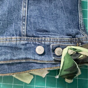 Puede incluir: Una chaqueta de mezclilla azul con una pretina que presenta dos botones met&aacute;licos grabados con el texto Old Navy. Una tira de tela de camuflaje en tonos verdes, marrones y negros est&aacute; anudada a trav&eacute;s del ojal en el lado derecho del dobladillo.