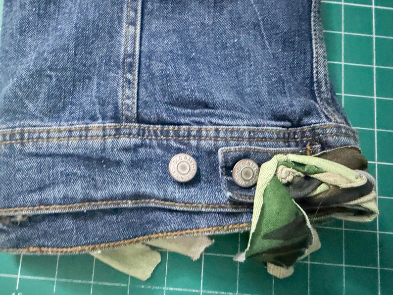 Puede incluir: Una chaqueta de mezclilla azul con una pretina que presenta dos botones met&aacute;licos grabados con el texto Old Navy. Una tira de tela de camuflaje en tonos verdes, marrones y negros est&aacute; anudada a trav&eacute;s del ojal en el lado derecho del dobladillo.