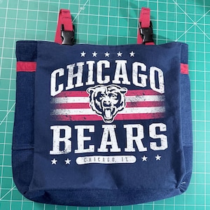 May include: A navy blue tote bag featuring the Chicago Bears logo with a bear graphic and the text "CHICAGO BEARS". The bag has red accents, denim sides, and red straps with black buckles. The text "CHICAGO, IL" is also visible.